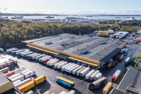 Flygfoto över en stor lagerlokal med flera lastbryggor och många lastbilar parkerade, omgiven av träd och ett industriellt hamnområde i bakgrunden.