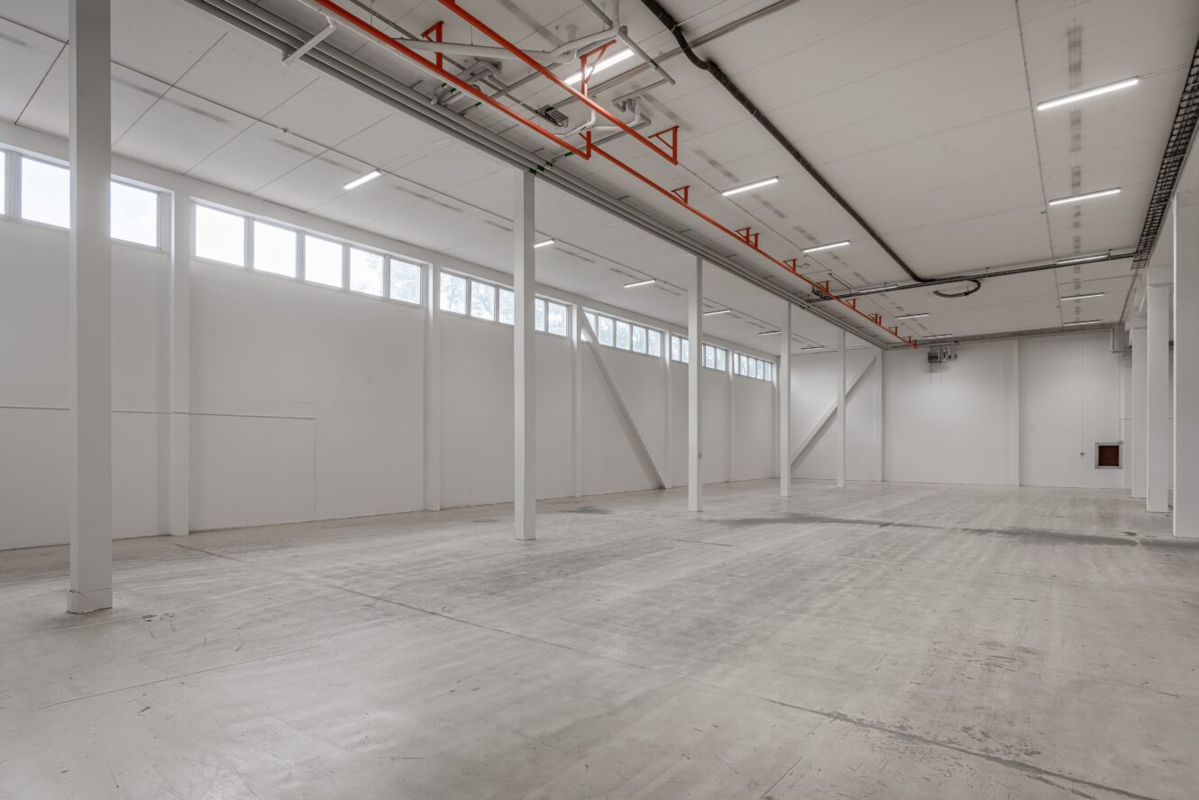 Rymlig, tom industrilokal med högt i tak, vita väggar, stora fönster samt synliga balkar och rör. Betonggolv täcker hela ytan.