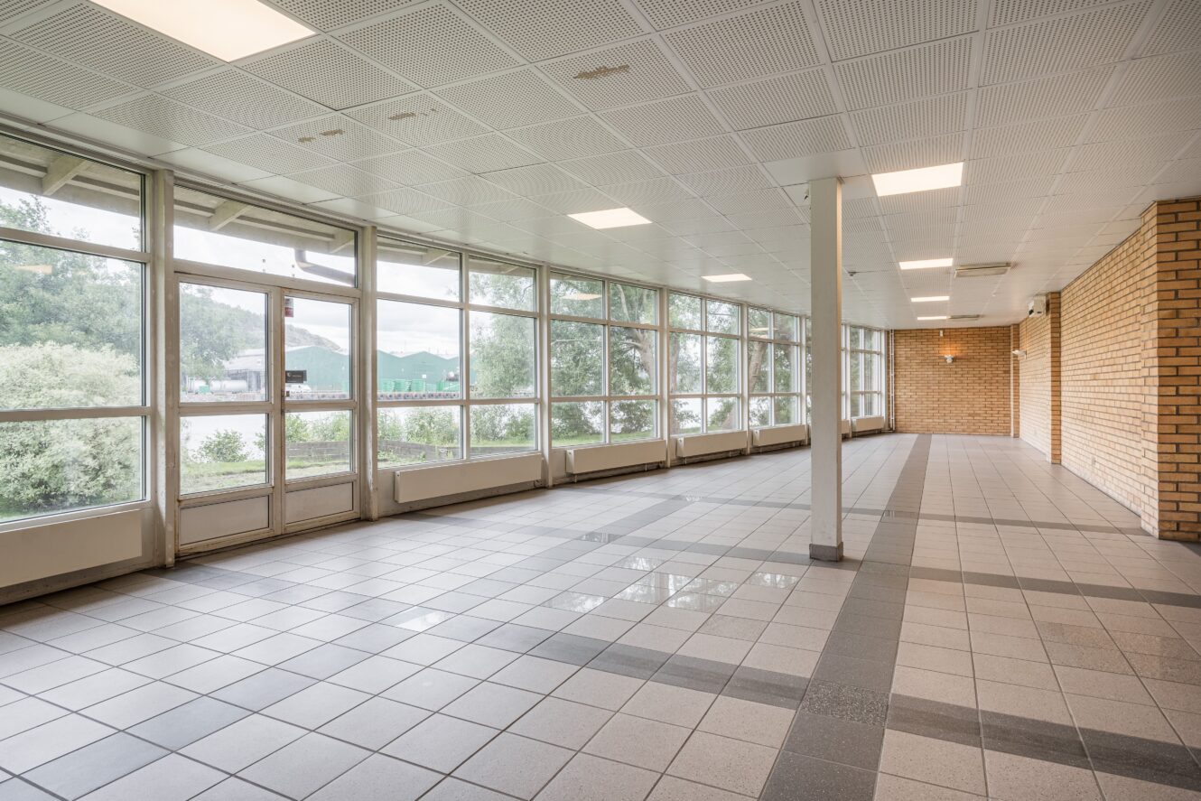 En tom hall med stora fönster, klinkergolv, tegelväggar och taklampor, med träd och grönska som syns utanför.