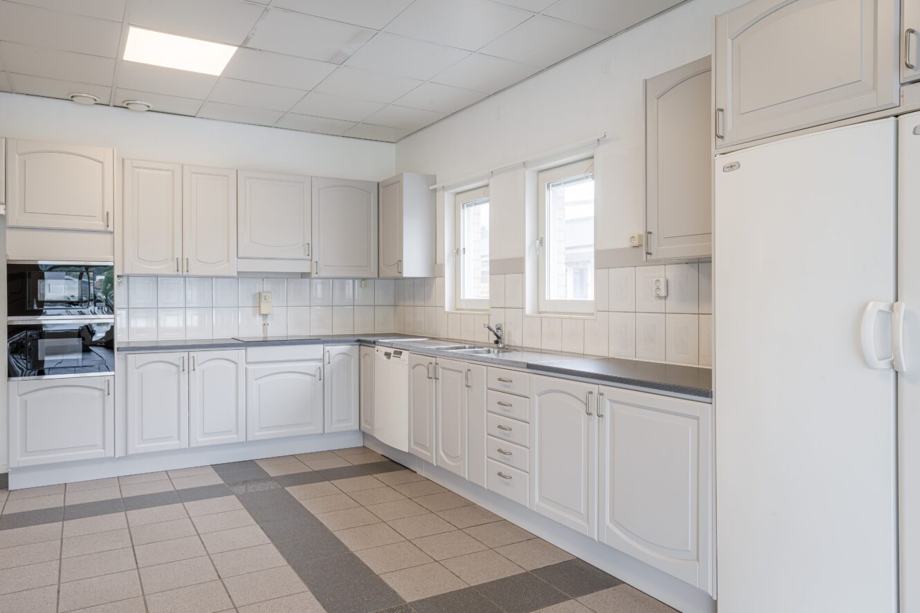 Rymligt kök med vita skåp, inbyggda vitvaror, kaklat stänkskydd, stort kylskåp och klinkergolv; två fönster ovanför diskbänken ger naturligt ljus.