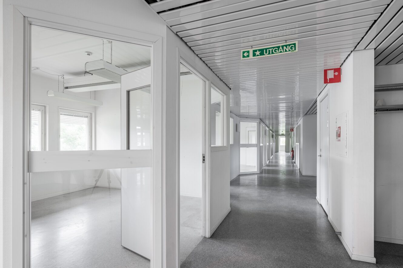 En ren, tom korridor med vita väggar och öppna dörrar som leder till lediga rum; en grön utgångsskylt med texten "Utgang" hänger från taket.