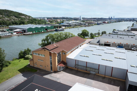 Industribyggnader och lagerlokaler ligger längs en flod, med en stadsbild och gröna kullar i bakgrunden under en delvis molnig himmel.