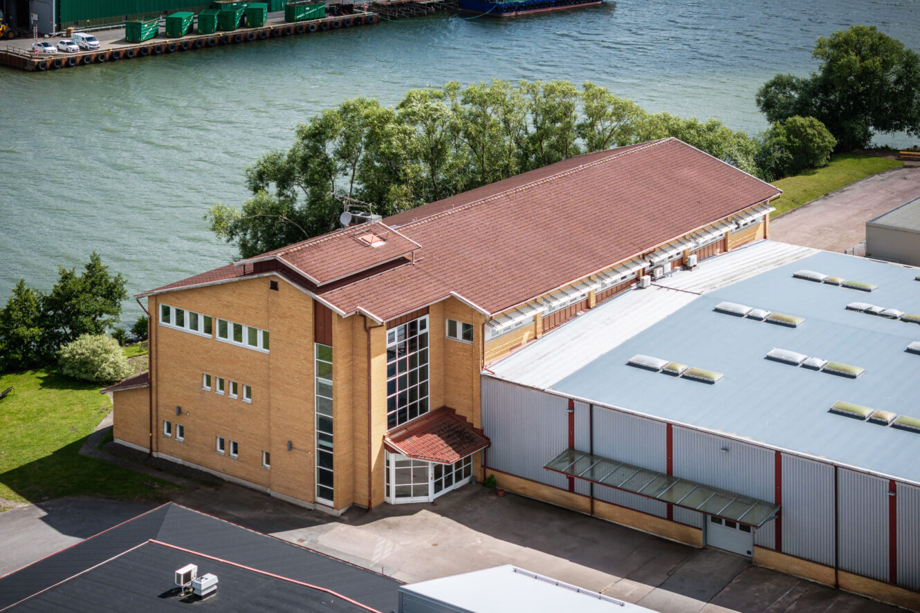 Flygfoto över en solbrun tegelbyggnad med rött tegeltak intill en stor lagerlokal i metall, belägen vid en flod och omgiven av träd.