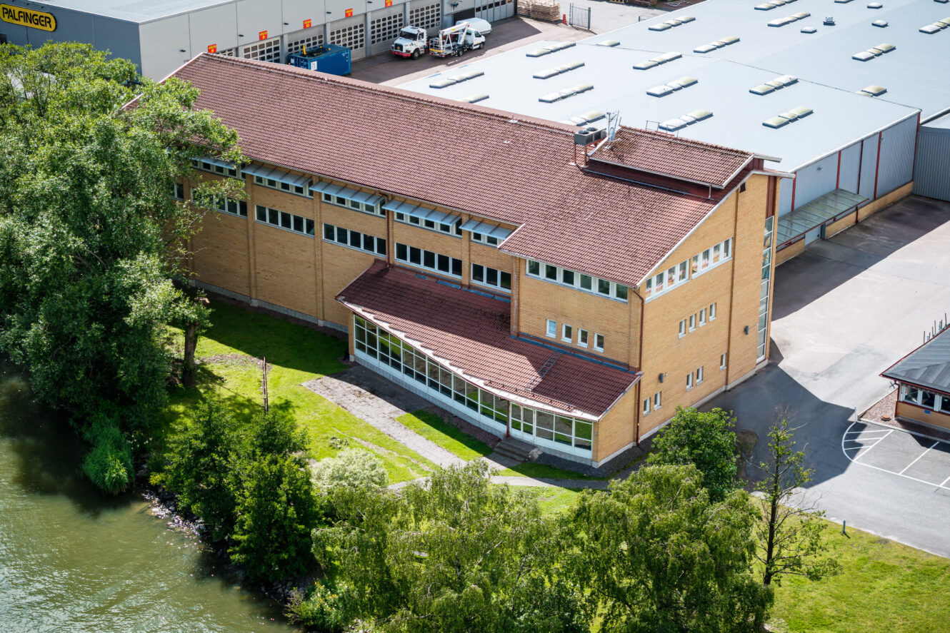 Flygfoto över en stor industribyggnad med gula tegelväggar och rött tak, belägen intill en flod och omgiven av träd och parkeringsplatser.