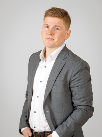 En man i grå kostym och vit skjorta står mot en ljus bakgrund och tittar in i kameran med ett neutralt uttryck.