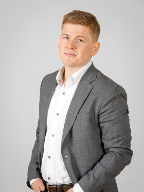 En man i grå kostym och vit skjorta står mot en ljus bakgrund och tittar in i kameran med ett neutralt uttryck.