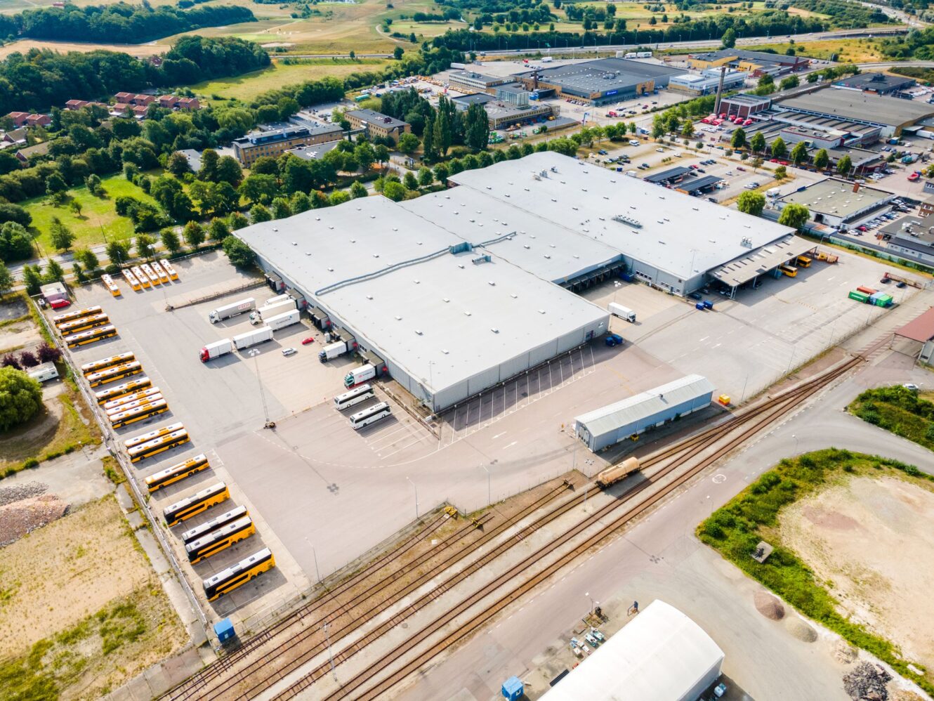 Flygfoto över ett stort lager eller distributionscenter med flera lastbryggor, parkerade bussar och intilliggande järnvägsspår i ett industriområde.