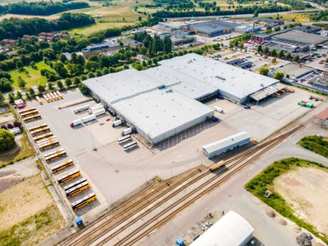 Flygfoto över ett stort lager eller distributionscenter med flera lastbryggor, parkerade bussar och intilliggande järnvägsspår i ett industriområde.