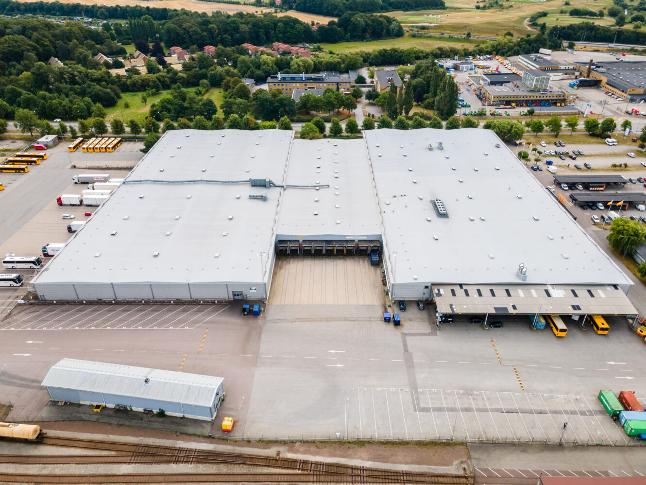 Flygfoto över ett stort industriellt lagerkomplex med flera lastbryggor, parkerade fordon och omgivande vägar och grönska.