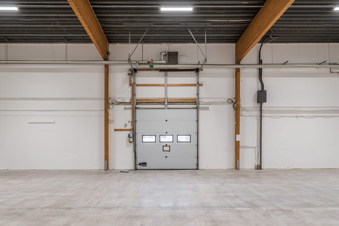 Tomt industriellt lagerutrymme med betonggolv, synliga takbjälkar och en stängd garageport mitt på bakväggen.