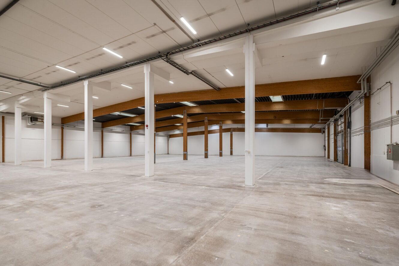 Rymlig, tom lagerlokal med synliga bjälkar, vita väggar, betonggolv och lysrörsbelysning.