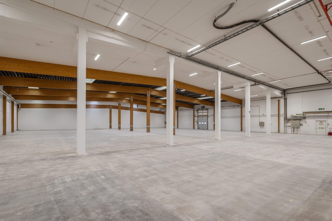 Rymlig, tom industrilagerinredning med betonggolv, vita väggar, synliga takbjälkar och takbelysning.