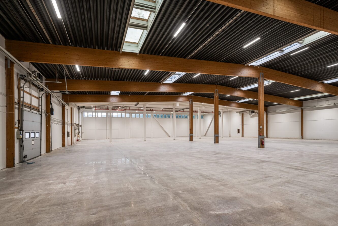 Stor tom lagerlokal med högt i tak, synliga träbjälkar, takfönster och betonggolv. Lysrör hänger från det mörka ribbade taket.
