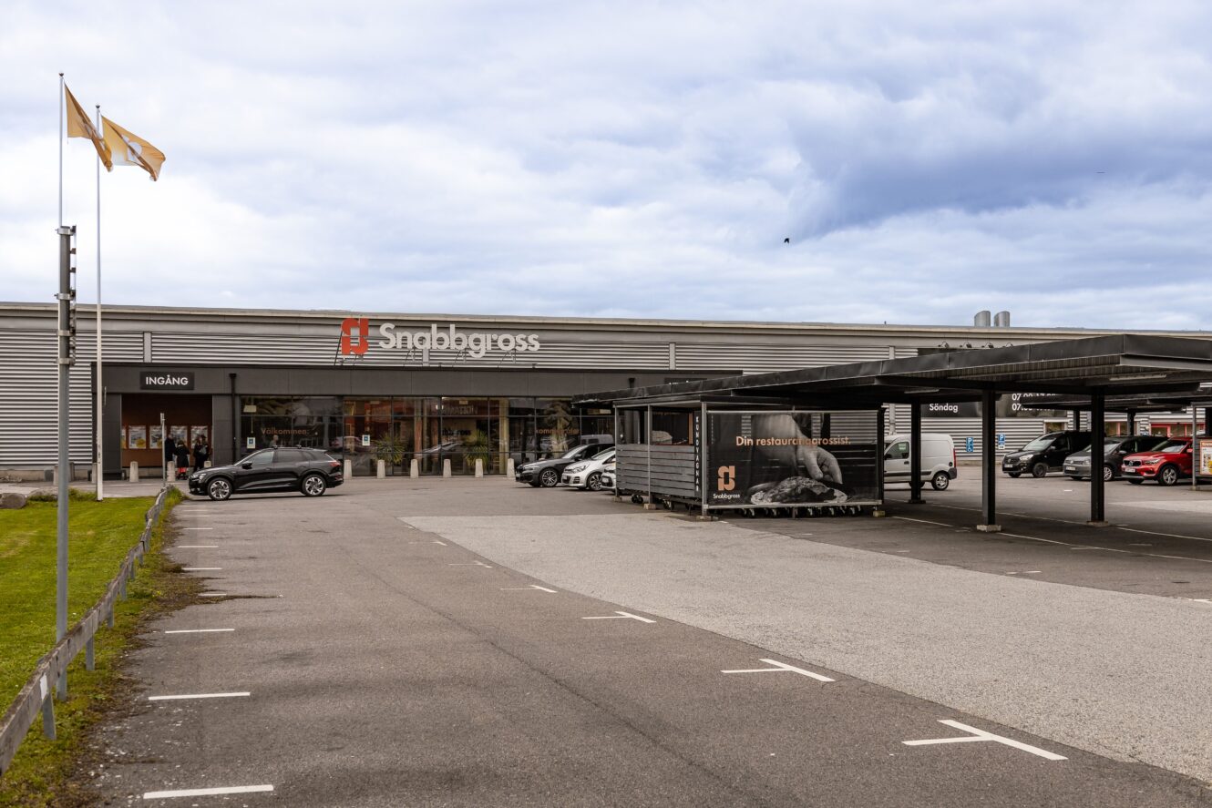 En stor Snabbgross-butik med parkeringsplats, flera parkerade bilar och vagnsförvaring under tak en molnig dag.