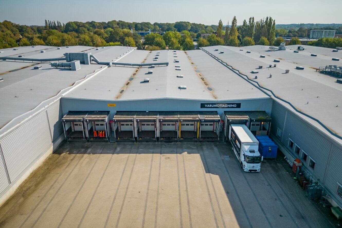 Flygfoto över en stor lagerlokal med flera lastkajer, en parkerad lastbil, blå containrar och träd i bakgrunden.