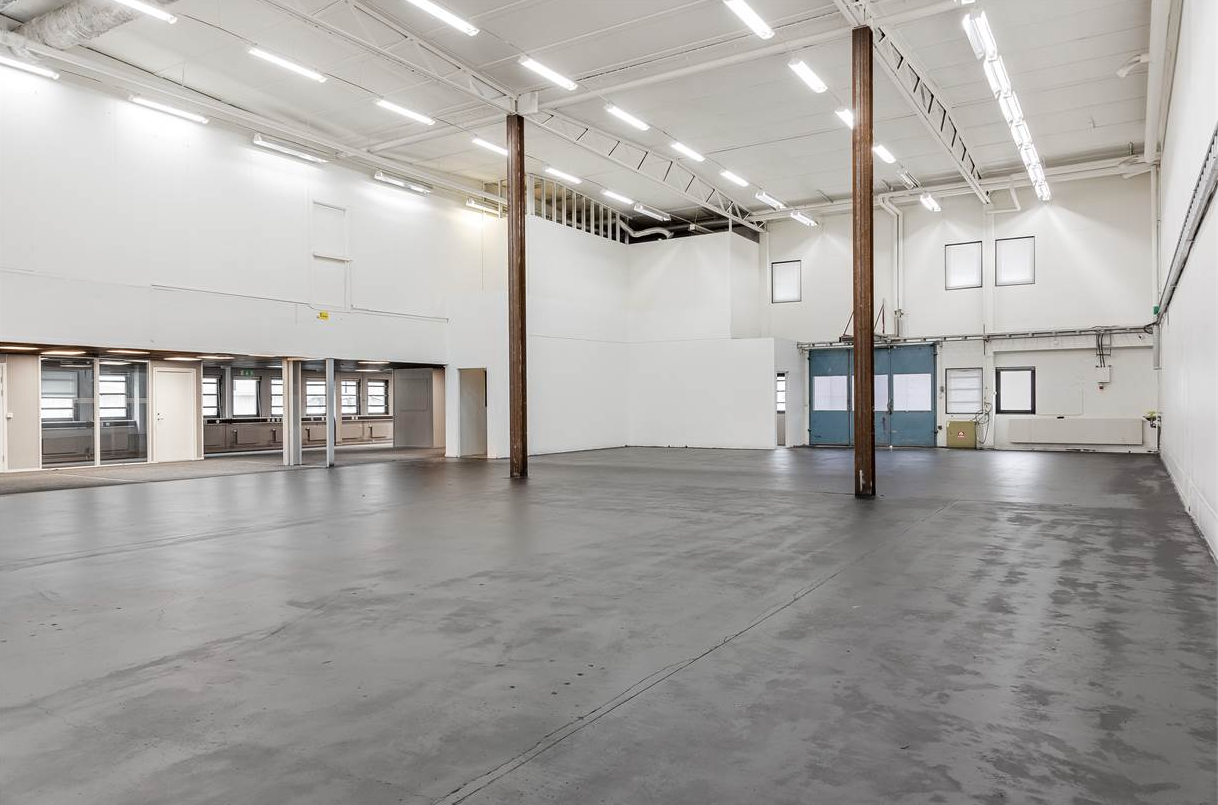 Rymlig, tom lagerlokal med högt i tak, vita väggar, stora fönster och polerade betonggolv, med synliga balkar och lysrörsbelysning.