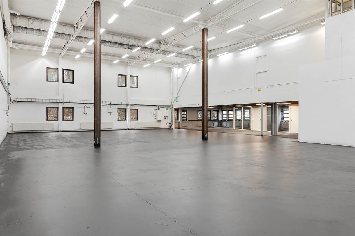 Rymlig, tom industrilagerlokal med vita väggar, högt i tak, synliga bjälkar och flera fönster och dörrar längs väggarna.