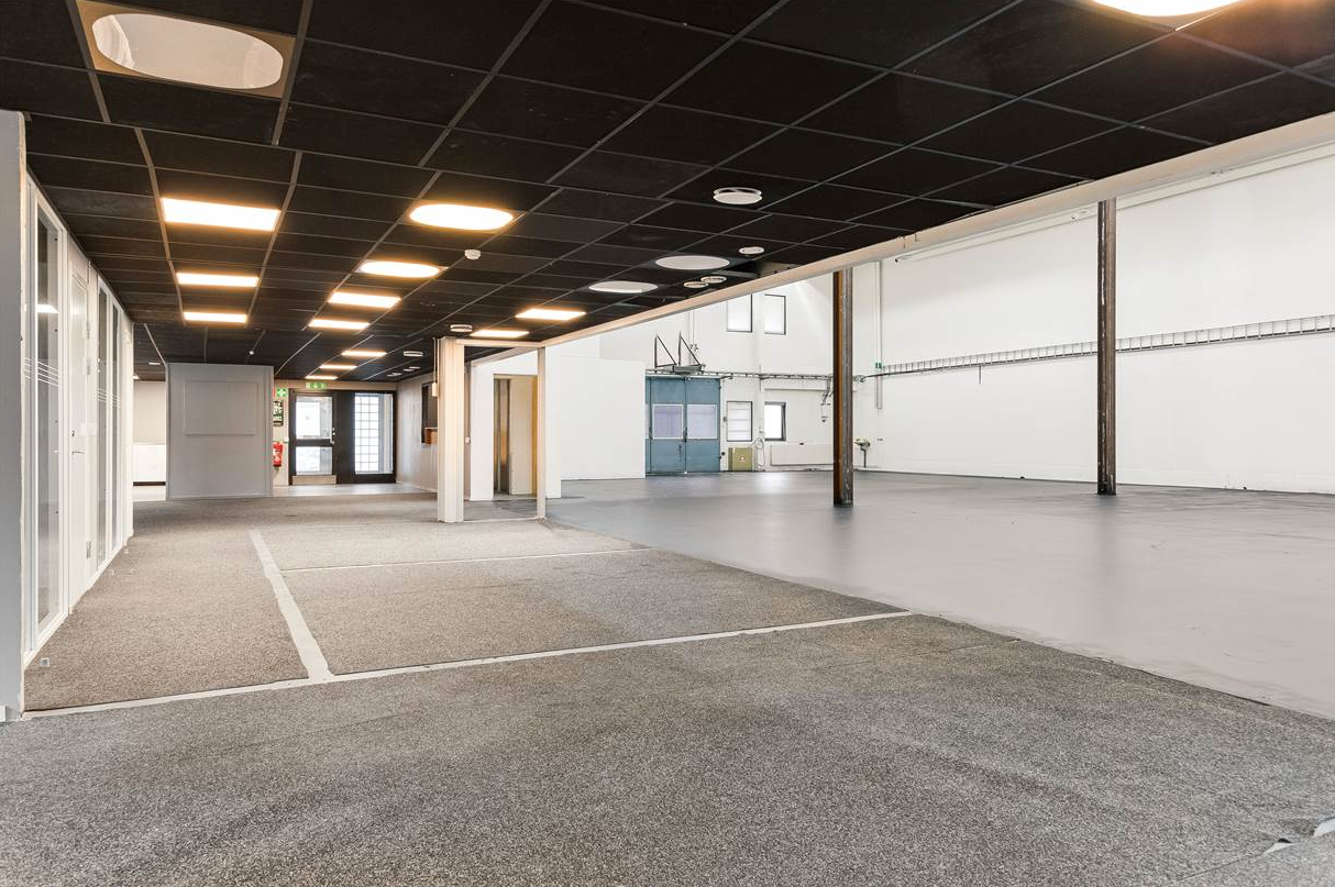 Rymlig, tom lokal med heltäckningsmatta och betonggolv, vita väggar, svart gallertak, synliga bjälkar och stora fönster.