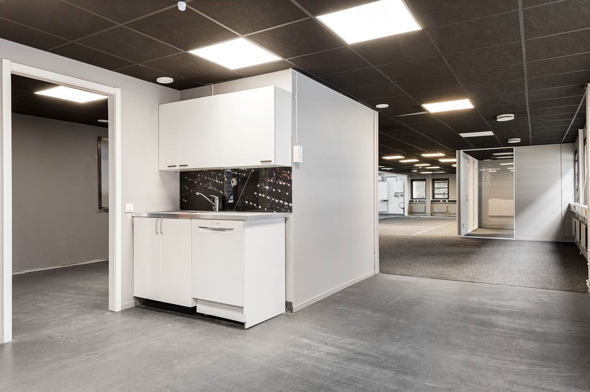 Kontorsinredning med ett litet pentry i förgrunden, grått golv, vita väggar, svarta takplattor och stark takbelysning.