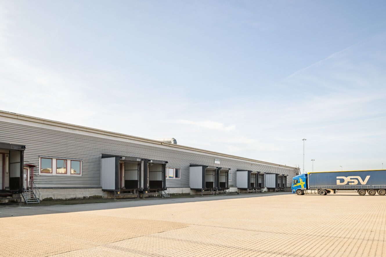 En stor lagerlokal med flera lastkajer och ett lastbilssläp med DSV-märke parkerat i närheten på en asfalterad tomt.