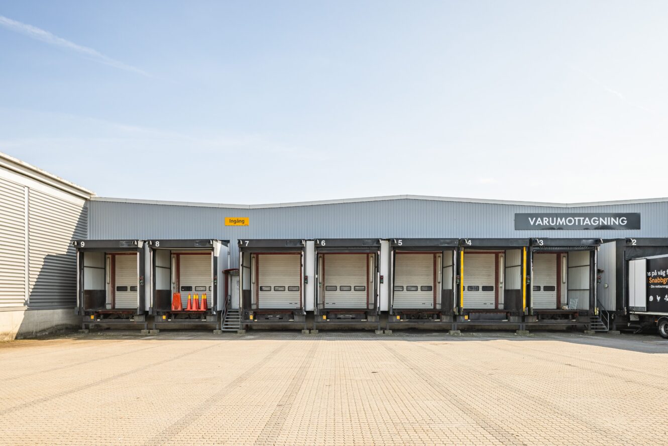 Bilden visar en rad med sex lastkajer med stängda dörrar på ett lager märkt VARUMOTTAGNING under en klar himmel.