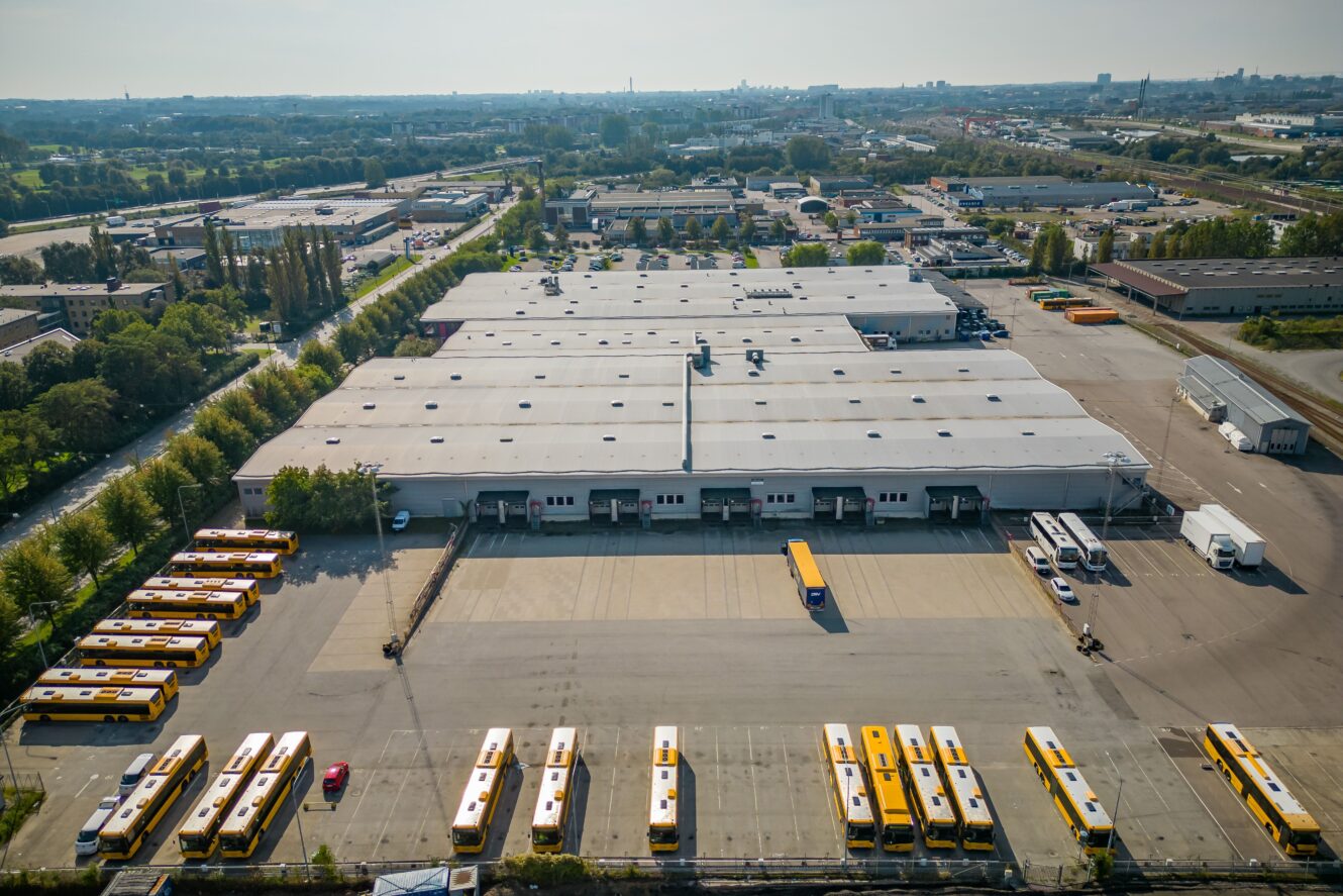 Flygfoto över en stor lagerlokal med flera gula bussar parkerade i rader runt byggnaden, omgiven av industrianläggningar och grönska.