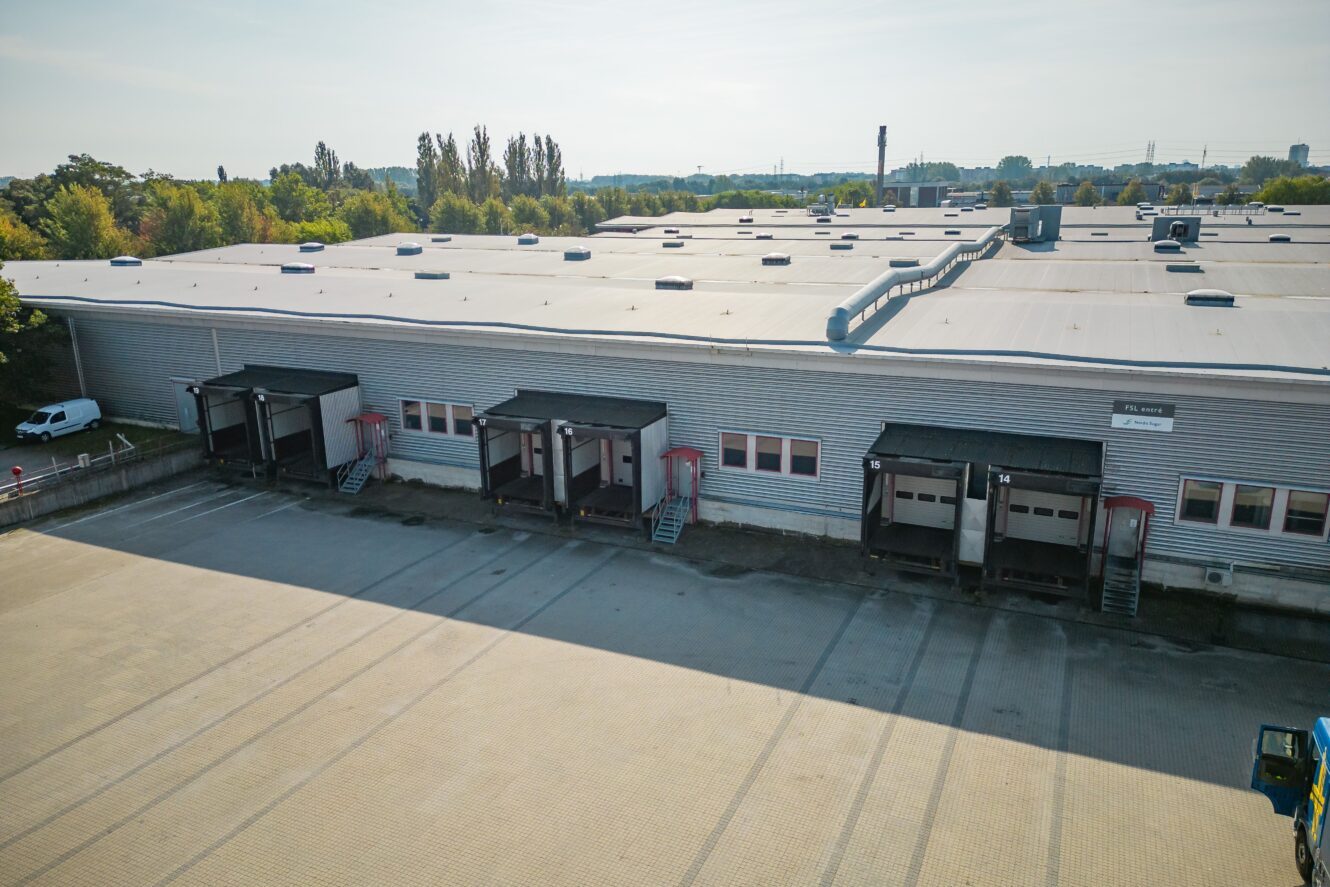 En stor industrihall med flera lastkajer och en tom asfalterad tomt framför, omgiven av träd under en klar himmel.