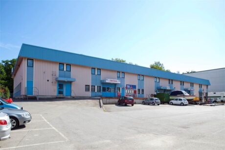 En lätt industribyggnad med blå dörrar och lister, en skylt ovanför entrén, flera parkerade bilar och en tom asfalterad tomt framför.