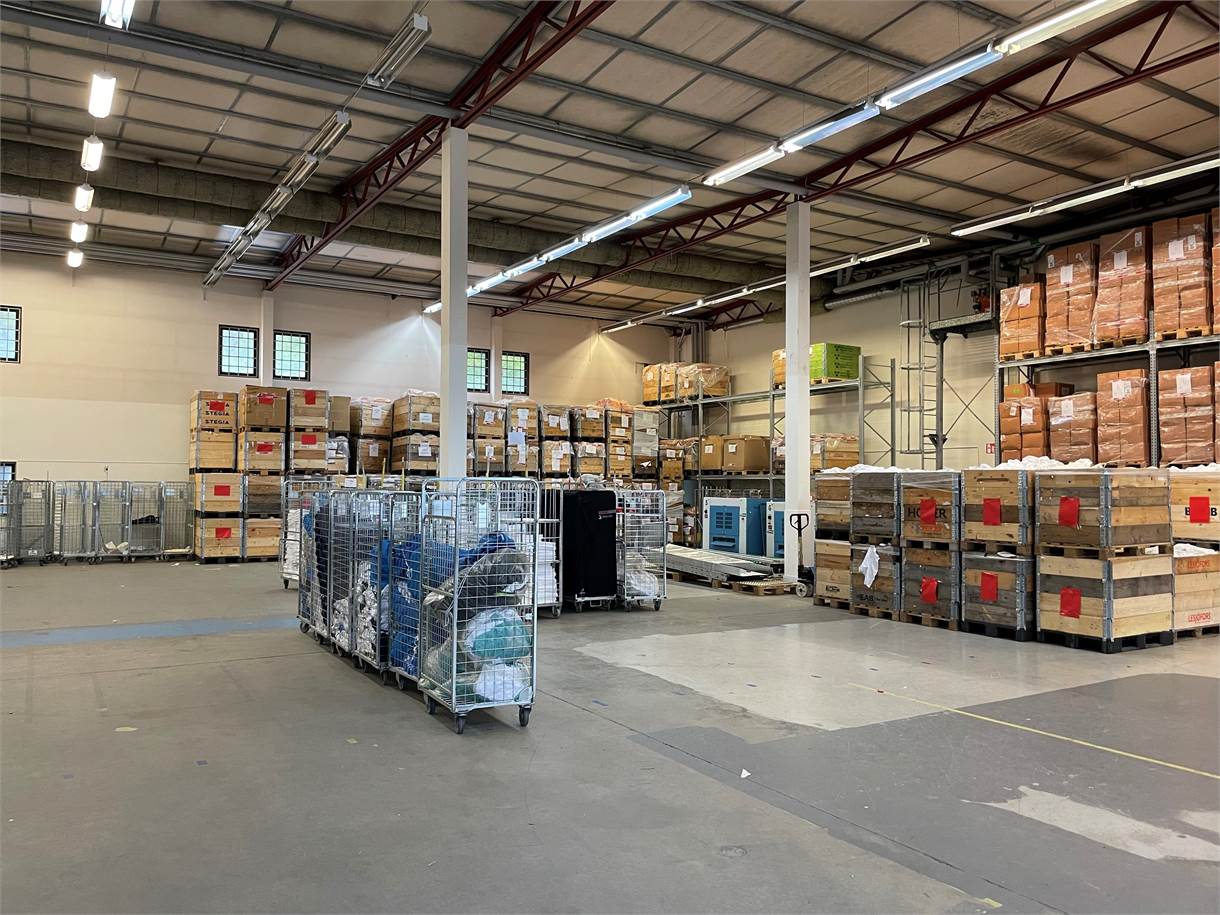 Stor lagerlokal med staplade pallar med lådor och förvaringsvagnar i metall som är placerade i hela det öppna utrymmet.