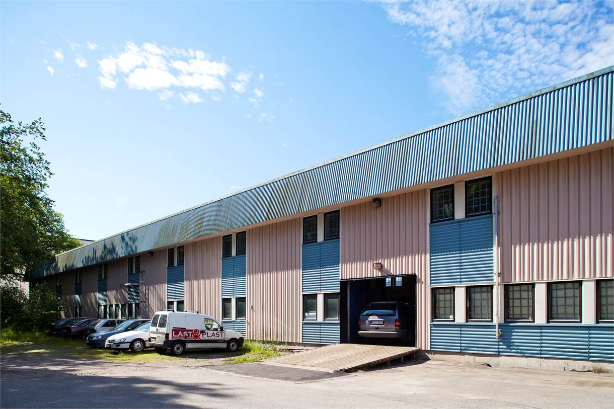 En stor industribyggnad med blå och beige metallfasad, parkerade bilar framför och en skåpbil märkt LASTPLAST vid entrén under en delvis molnig himmel.