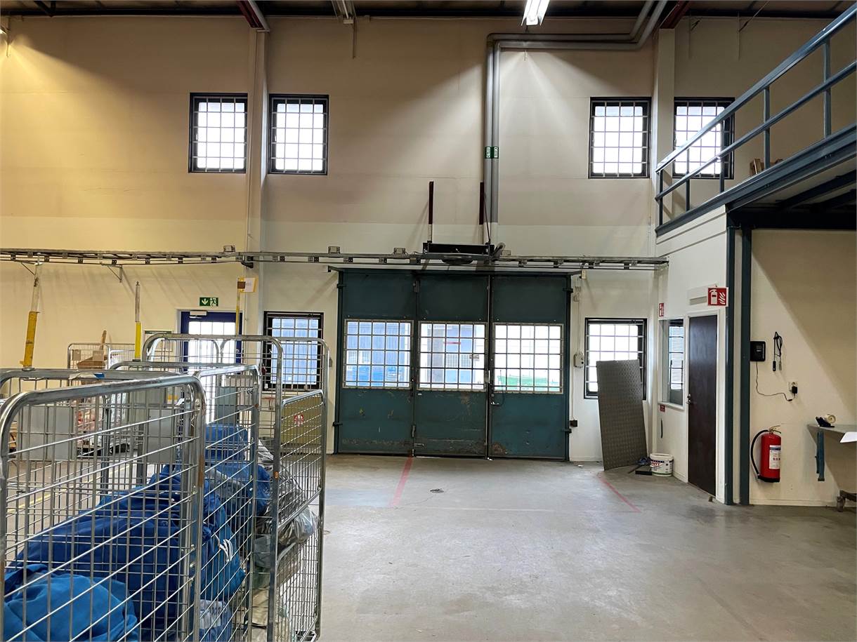 Industriell lagerlokal med lagervagnar i metall, en stor stängd lastport, rörledningar och synlig säkerhetsutrustning på väggarna.