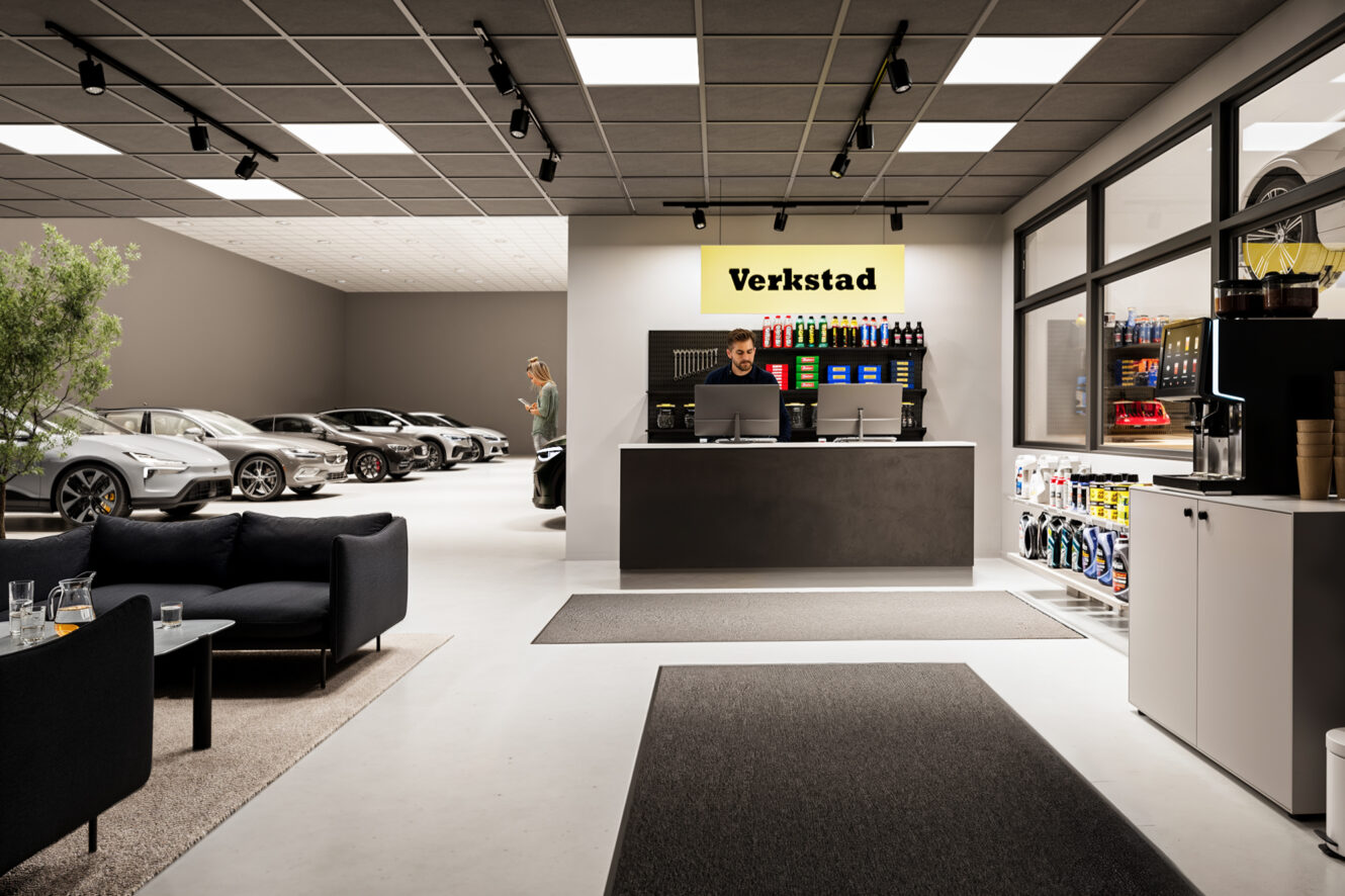 Modern bilverkstadsreception med reception, receptionist, väntrum och flera bilar i serviceområdet; hyllor med bilprodukter.