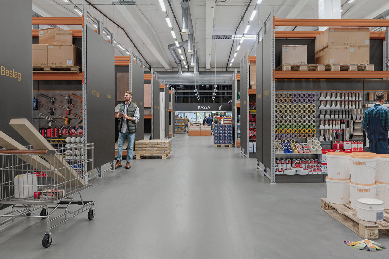 En järnhandel med hyllor med verktyg, färgburkar och förnödenheter; en man skjuter en kundvagn medan en annan handlar och en kassaskylt visar till kassan längre fram.