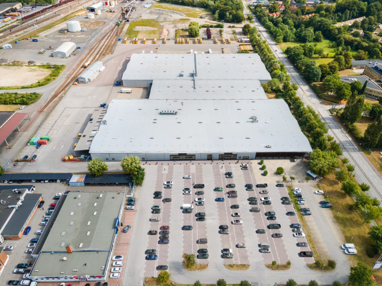 Flygfoto över en stor lagerbyggnad med en parkeringsplats full av bilar framför, omgiven av vägar, träd och industriområden.