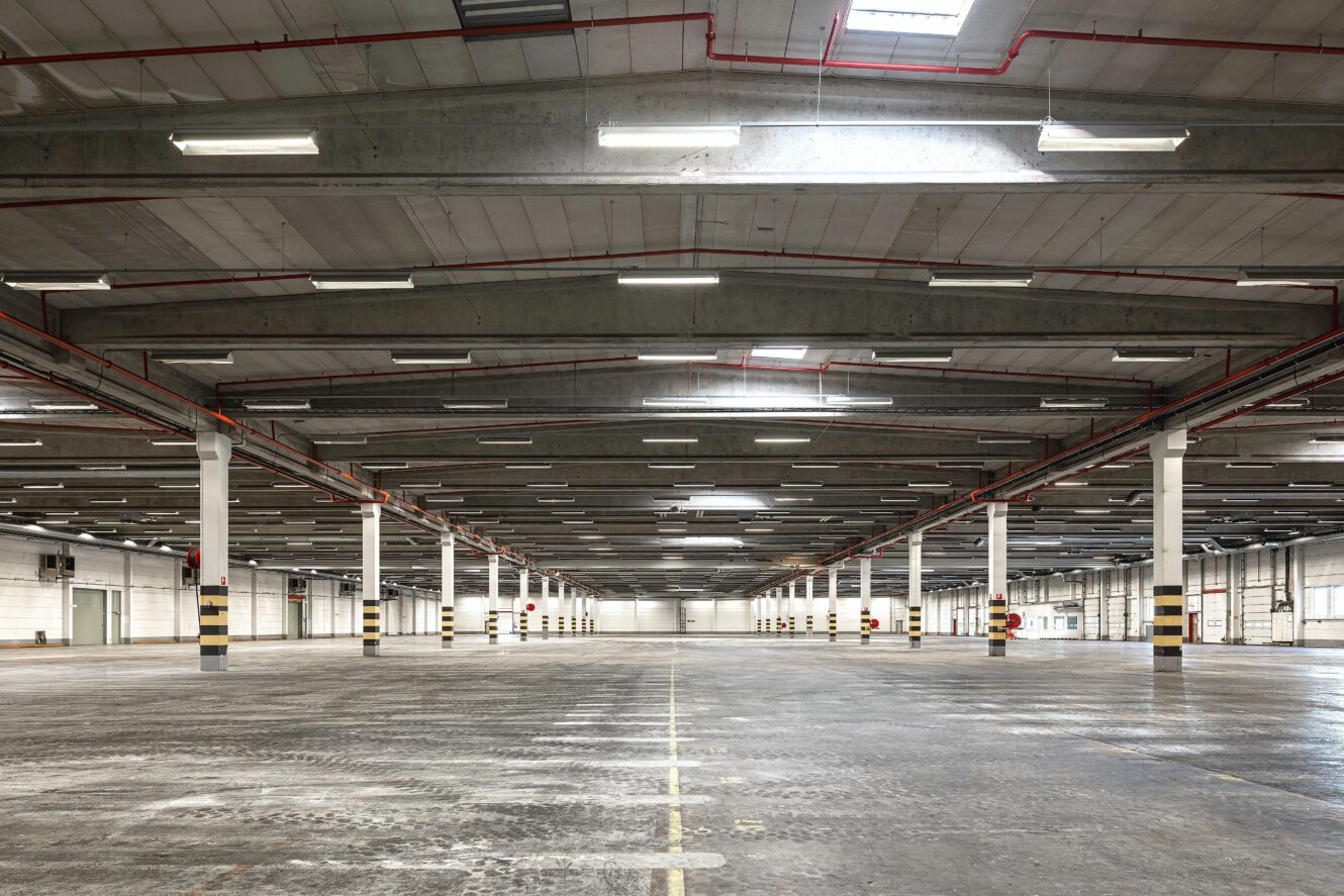Stor, tom lagerlokal med betonggolv, högt i tak, synliga rör och jämnt fördelade vita stödpelare under fluorescerande belysning.