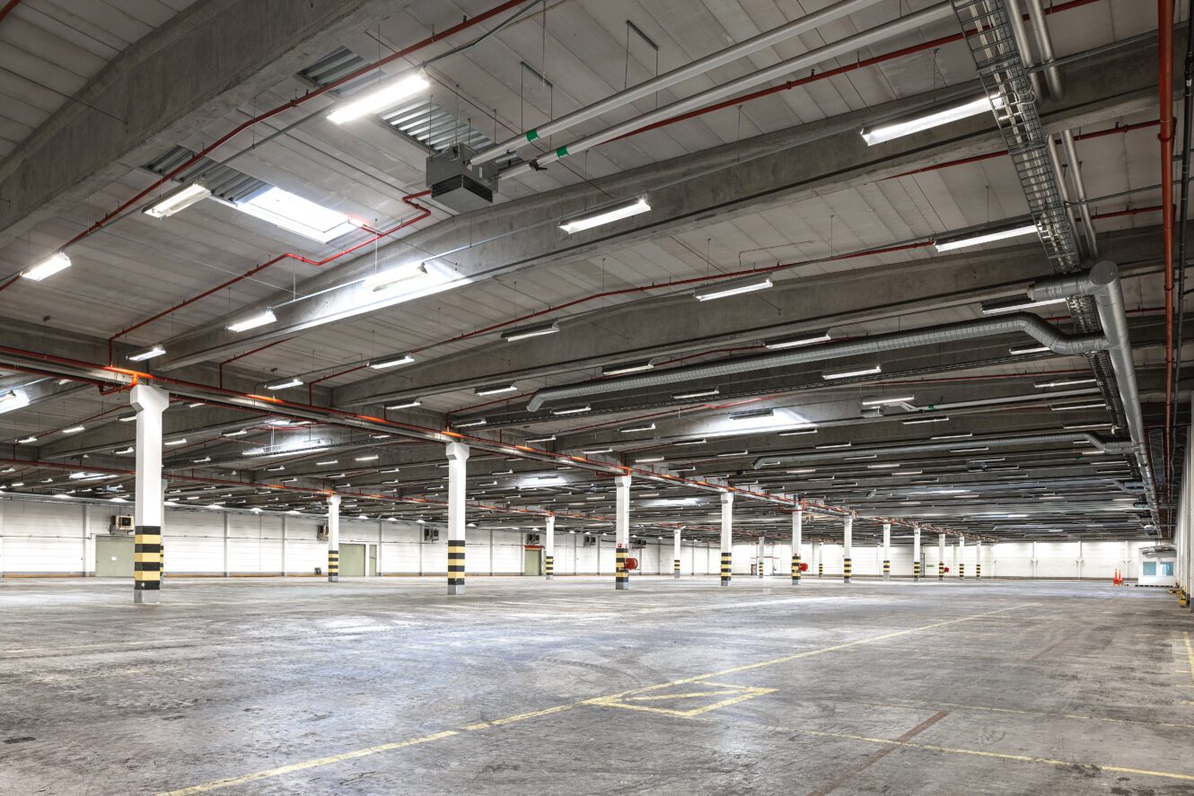 Stor, tom industrilagerlokal med betonggolv, högt i tak, stödpelare, takbelysning och synliga rör och kanaler. Gula och svarta markeringar på pelare och golv.