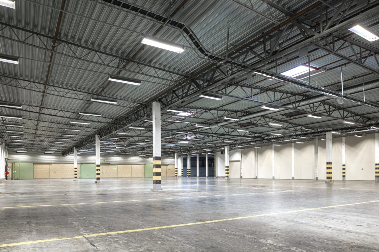Stor tom lagerlokal med betonggolv, synliga takbjälkar, takbelysning och markerade parkeringslinjer.