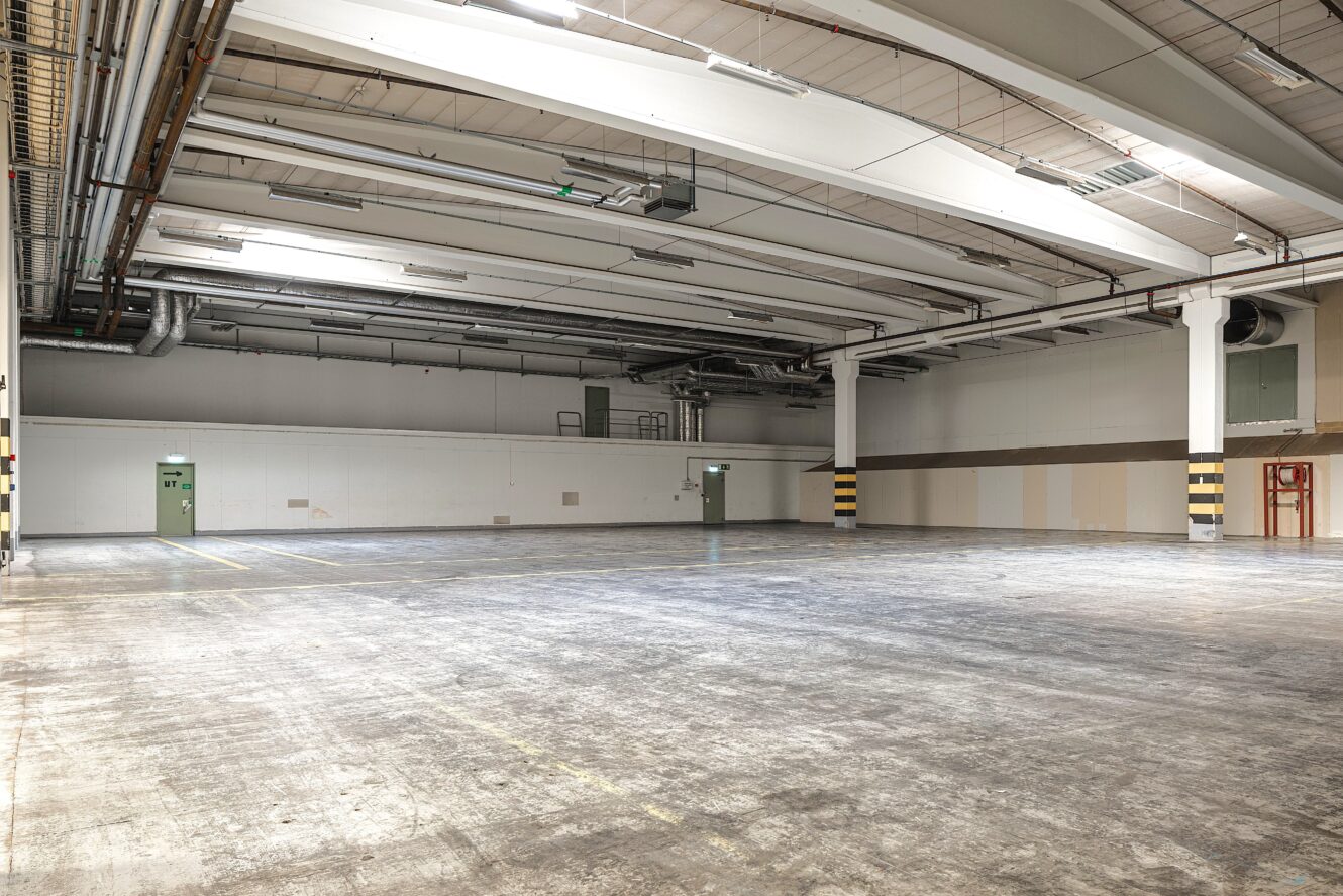 Stort tomt industriellt lagerutrymme med betonggolv, takbelysning, synliga rör och två gröna utgångsdörrar på den bortre väggen.