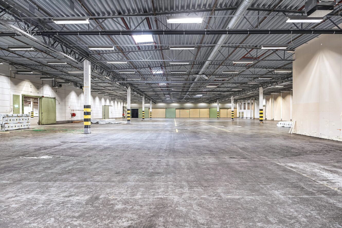 Stort tomt industrilager med betonggolv, synliga rör i taket och jämnt fördelade stödpelare. Starka taklampor och vita väggar är synliga i hela utrymmet.