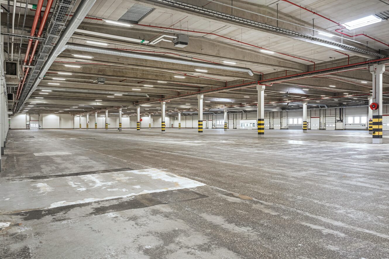 Stort, tomt industrilager med betonggolv, vita pelare, synliga tak och takbelysning.