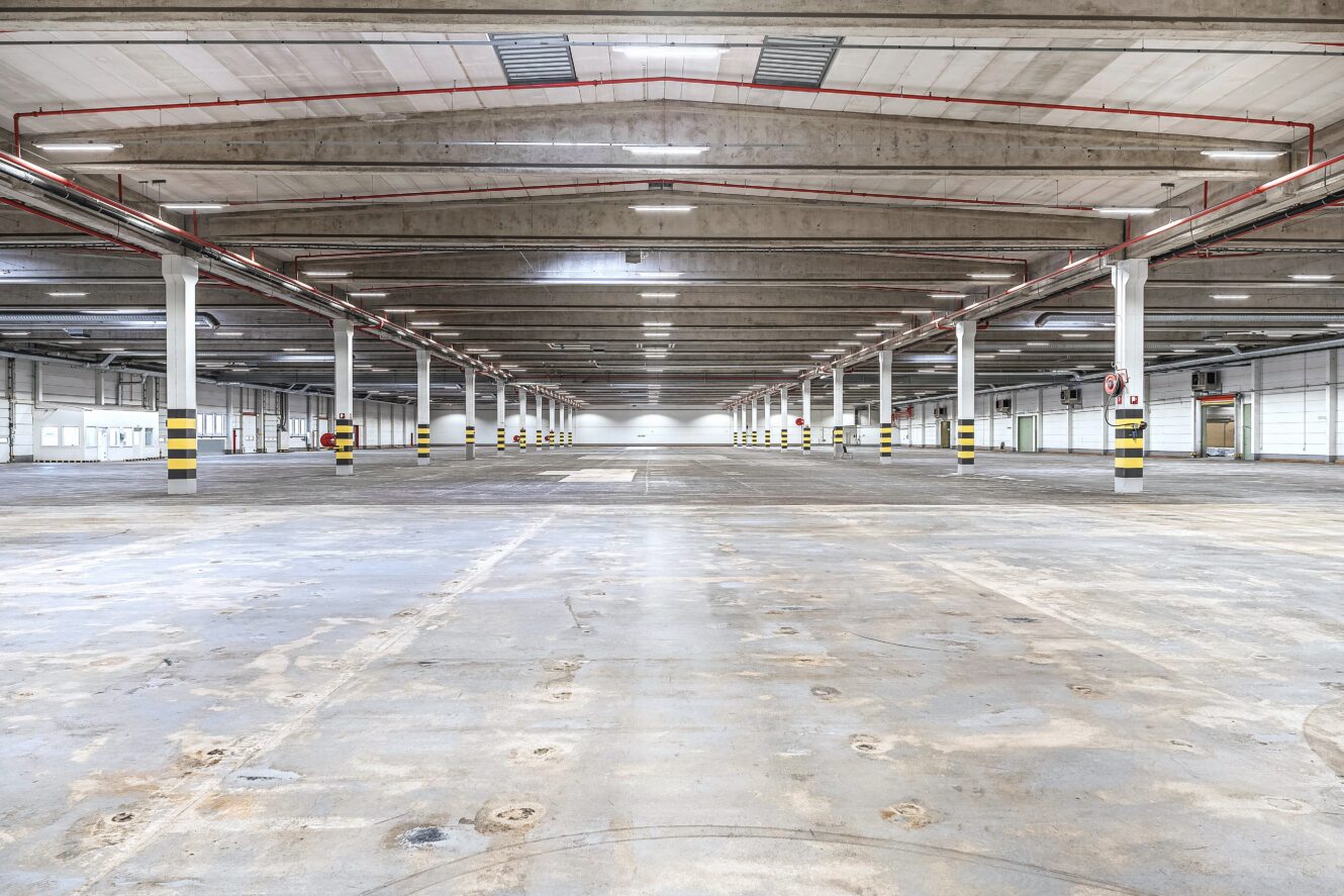Interiör av en tom industrilagerlokal med betonggolv, högt i tak, stödpelare, takbelysning och synliga ventilationskanaler.