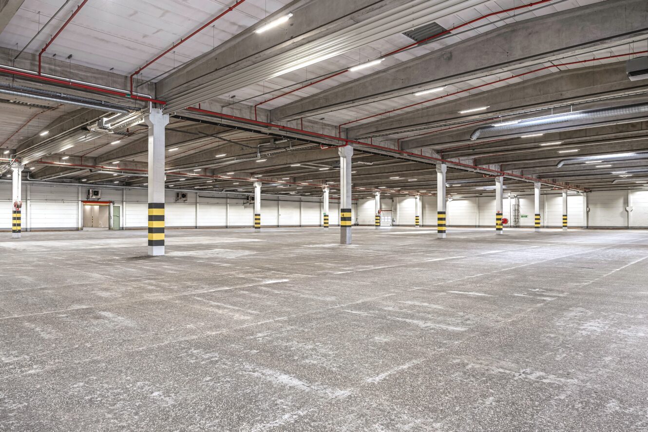 Stort, tomt industrilager med exponerade betonggolv, pelare och takbjälkar, lysrörsbelysning och synliga rör och ventiler.