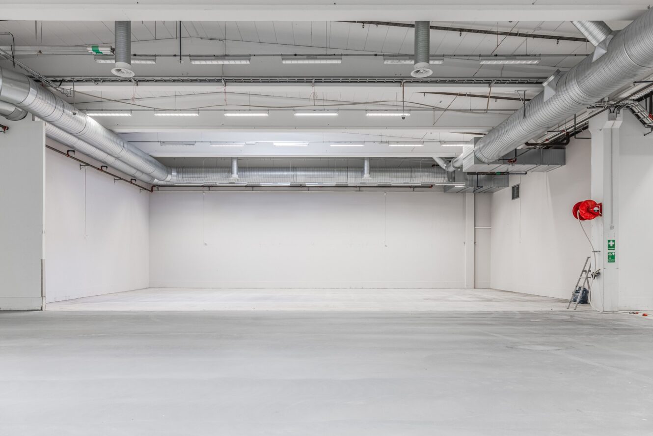 Tomt industrilager med vita väggar, synliga ventilationskanaler, starka taklampor och en brandslangsrulle på den högra väggen.