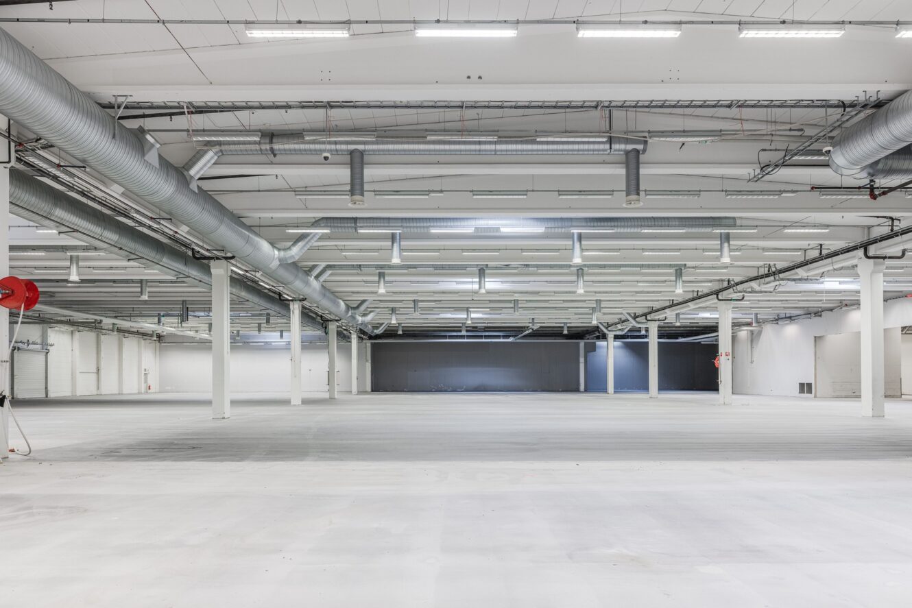 Stor tom lagerlokal med vita väggar, högt i tak, synliga kanalsystem, takbelysning, betonggolv och stödpelare.