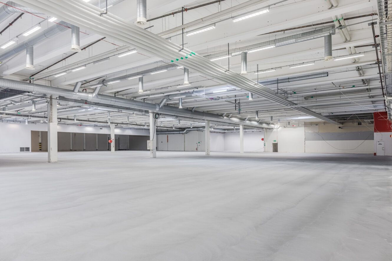 Stort tomt industrilager med vita väggar, högt i tak, synliga kanaler, takbelysning och betonggolv.