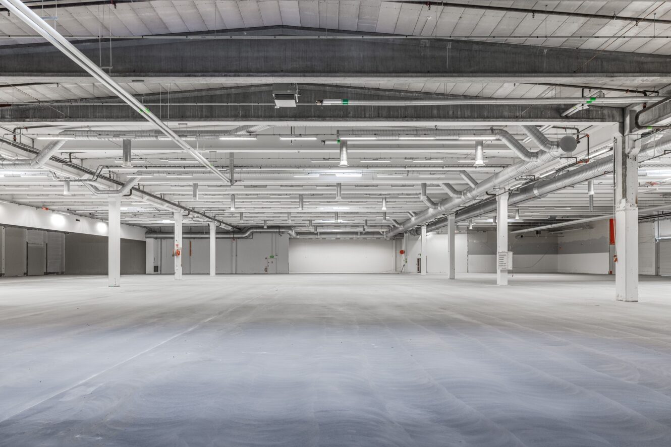 Stor, tom industrilagerlokal med synliga kanaler, vita väggar, stödpelare och betonggolv som lyses upp av taklampor.