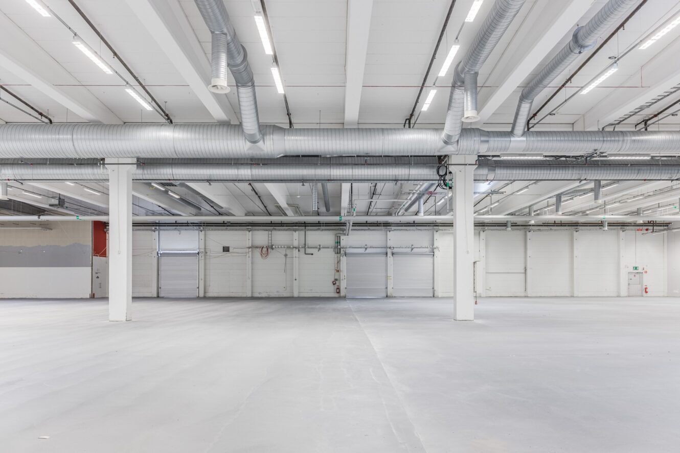 Stort tomt industrilager med vita väggar, grå golv, takbelysning och synliga ventilationskanaler i taket.