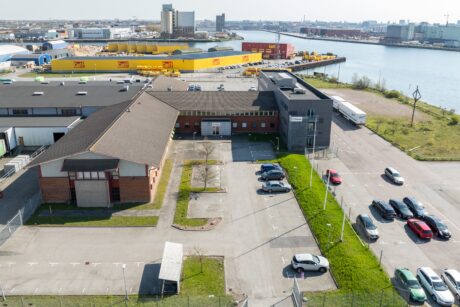 Flygfoto över en industribyggnad med en parkeringsplats framför, flera parkerade bilar och en flod med containrar och lagerlokaler i bakgrunden.