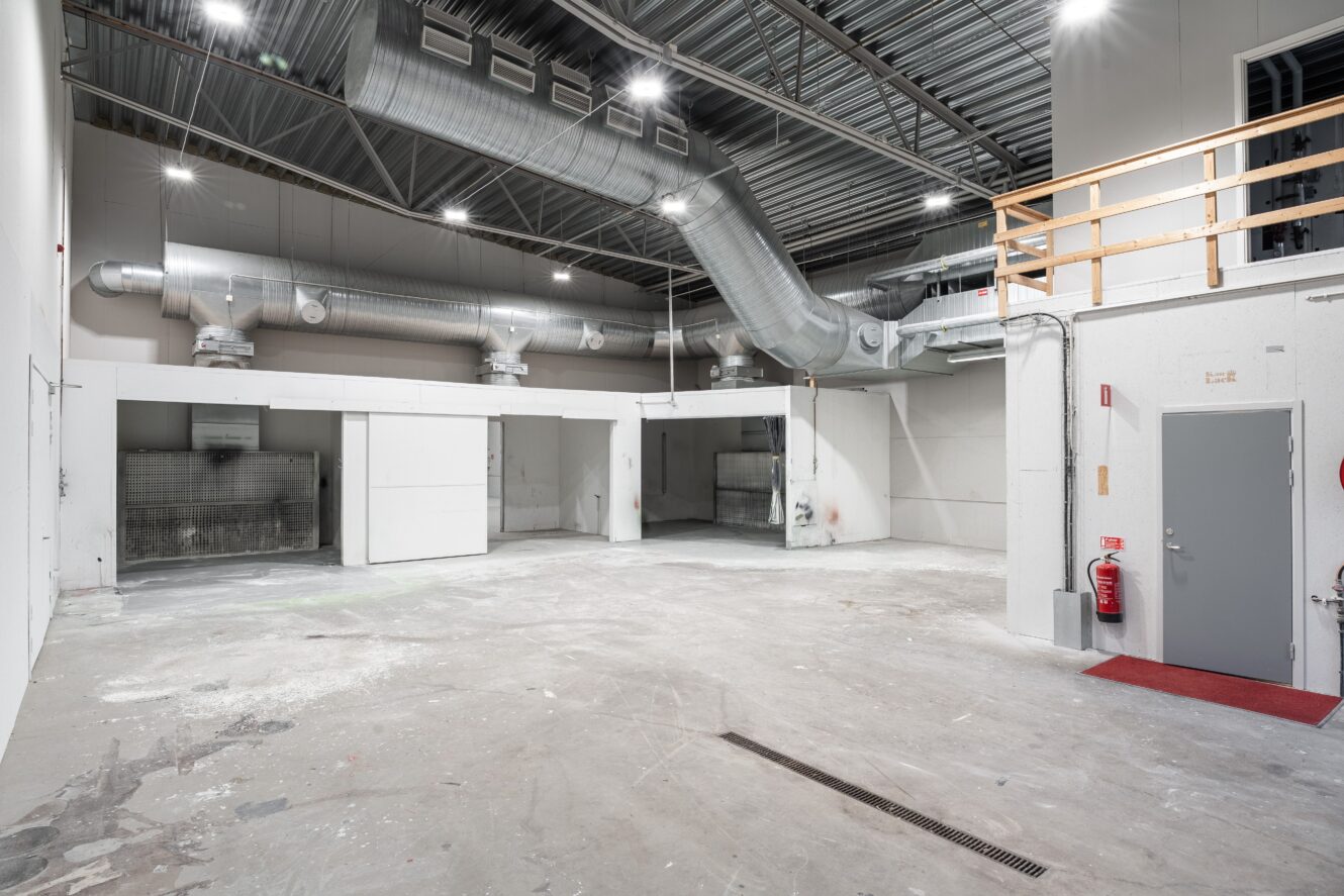 Industriell interiör med synliga ventilationskanaler, betonggolv och vita skiljeväggar; metalltak och ett entresolplan med träräcke är synliga.