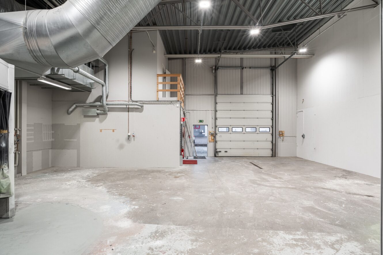 Tomt industrilager med högt i tak, synliga kanaler, betonggolv och en stor garageport.